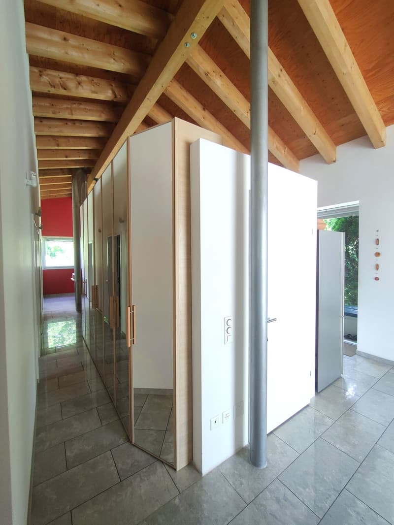 The image shows the interior of a building with a wooden ceiling and beams, white walls, and a tiled floor. There are several white cabinets or closets lining the hallway, and a large window at the end of the hallway providing natural light.