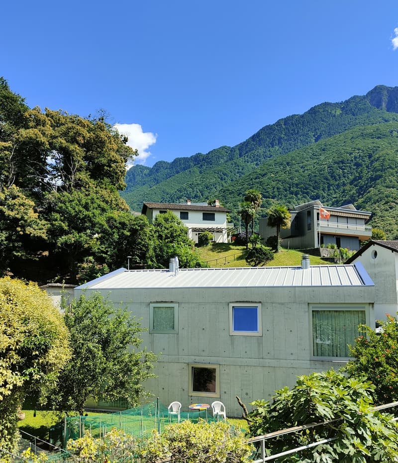 Two-story house with a green exterior, surrounded by lush greenery and mountains in the background. The house has a balcony or terrace visible.
