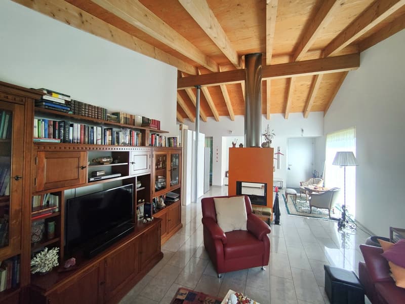 Spacious living room with exposed wooden beams on the ceiling, a large wooden bookshelf unit, a red leather armchair, and a fireplace in the corner. The room has tile flooring and large windows providing natural light.
