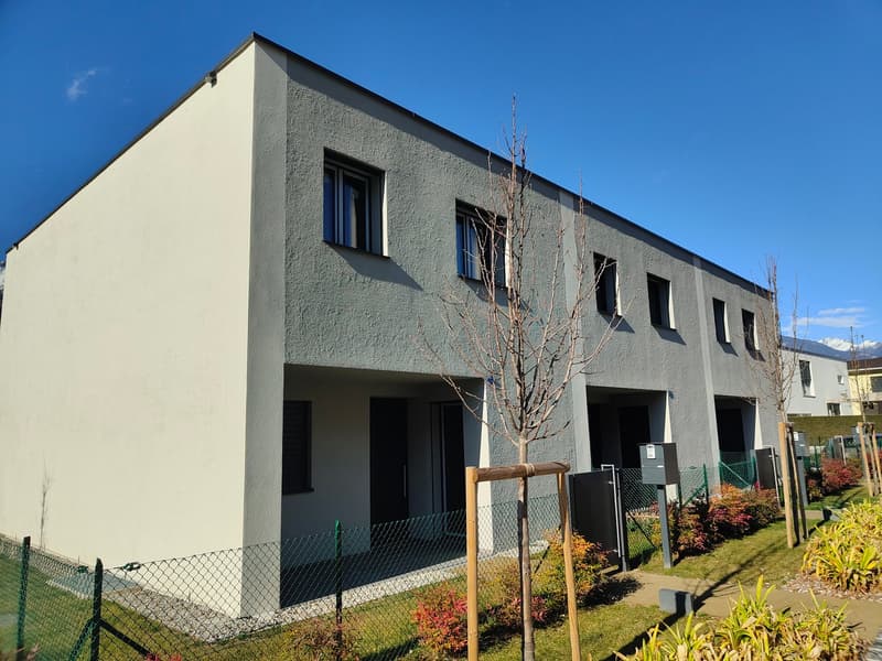 Modern multi-story building with gray walls, several windows, a fenced area, a small tree, and a paved pathway.
