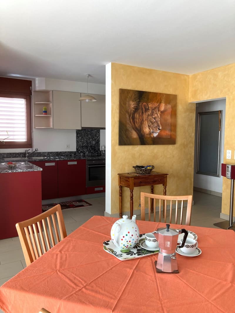 dining room with dining table, chairs, coffee maker, kettle, cups, plates, table runner, kitchen with red cabinets, counter top, sink, oven, lion painting on wall