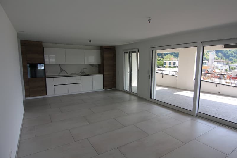 Spacious, open-plan room, tile flooring, large sliding glass doors leading to balcony, modern kitchen with white cabinets and dark wood accents, under-counter appliances.
