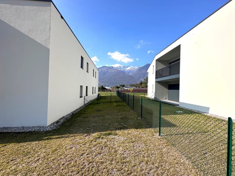 The image shows a multi-story apartment complex with white exterior walls and balconies. There is a grassy area with a fence in the foreground, and in the background, there are snow-capped mountains visible, indicating a scenic, mountainous location.