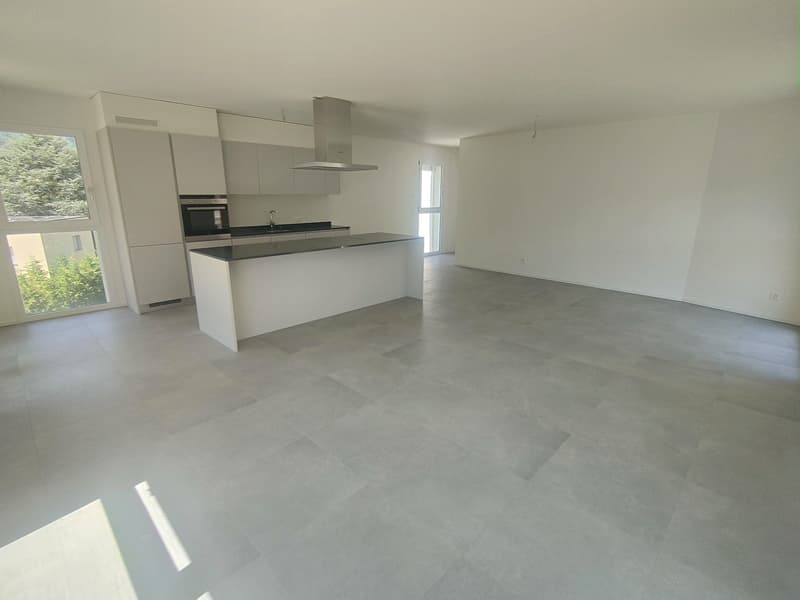 Open-concept living space with a modern kitchen featuring white cabinets, a stainless steel range hood, and a dishwasher. The flooring is a light-colored tile or laminate. Large windows provide natural light and a view of the surrounding area.