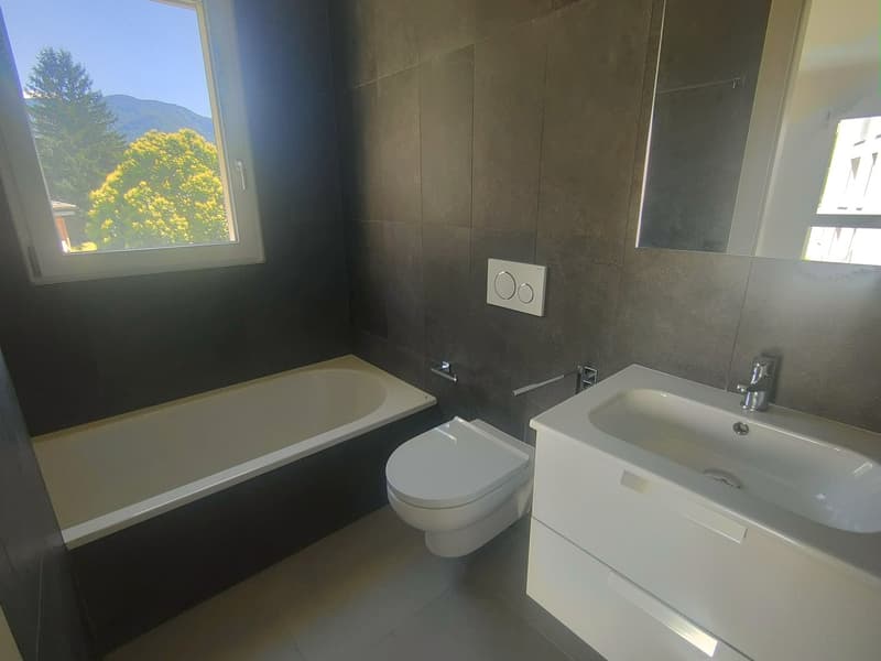 The image shows a modern bathroom with a bathtub, toilet, and sink. The walls are tiled in a dark color, and there is a large window providing natural light. The bathroom appears to be well-designed and equipped with contemporary fixtures.