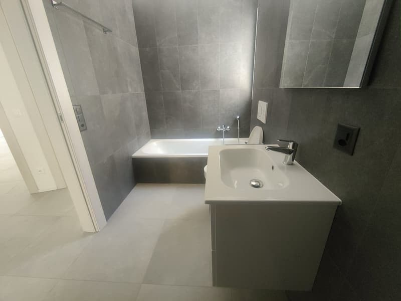 Large bathroom with grey tile walls, a white bathtub, a white sink vanity, and a mirror on the wall.