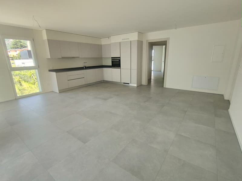 The image shows a modern, open-plan apartment with a spacious kitchen and living area. The kitchen features white cabinets and drawers, a black countertop, and built-in appliances. The flooring throughout the apartment is a light-colored tile or laminate