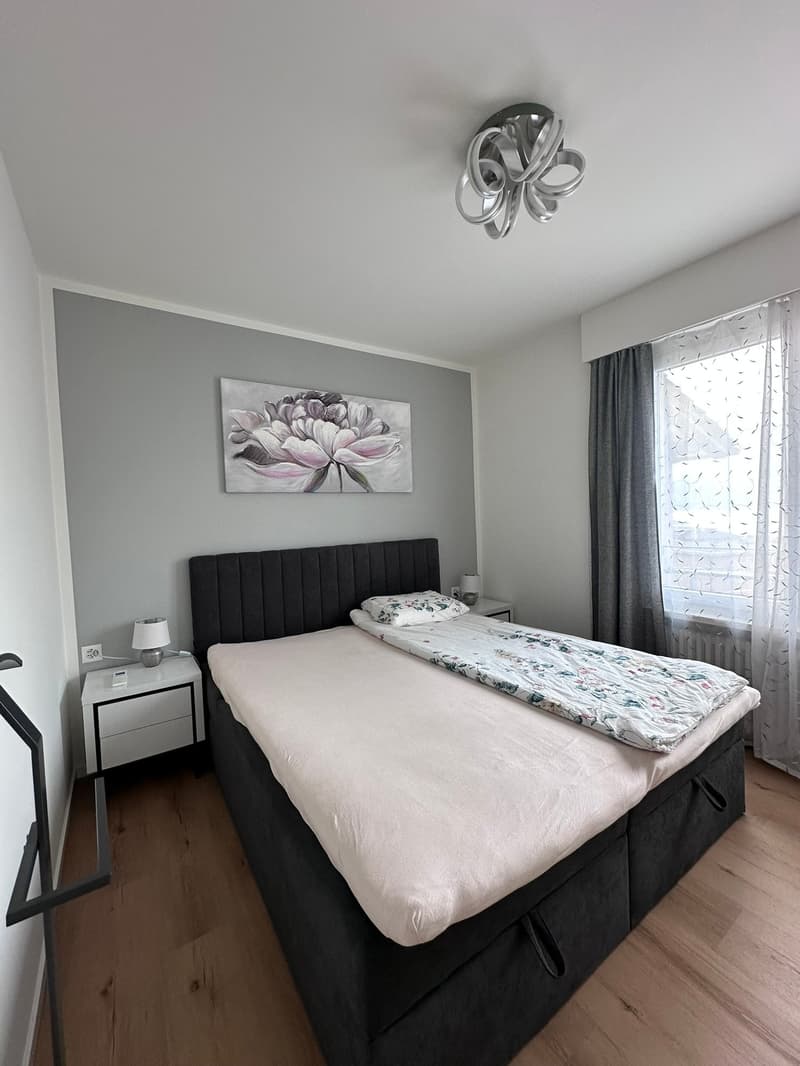 A bedroom with a double bed, nightstands, lamps, and a wall art. The bed has a pillow and a blanket. The wall has a mounted power outlet and a mounted picture frame. There is a glass window with curtains. The room has a wooden floor and white walls.