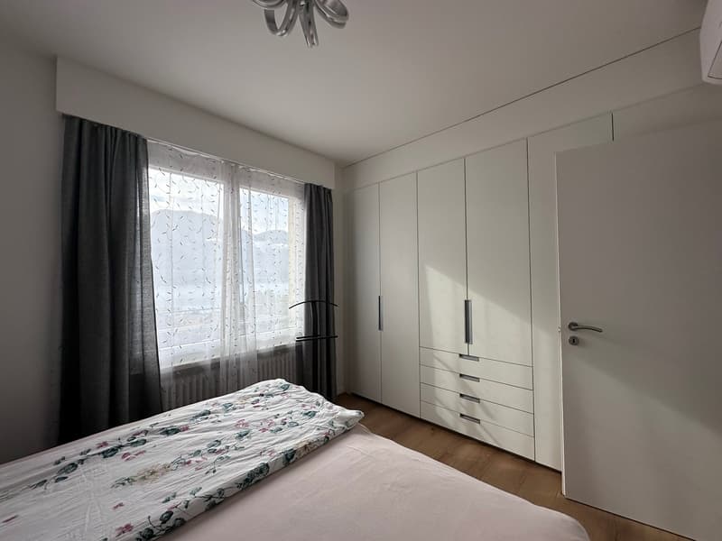 Bedroom with white walls, bed, white curtains, wardrobe, floor-mounted radiator, window with mountain view.