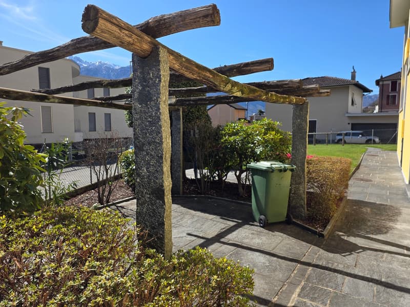 Outdoor area with wooden pergola, stone pillars, green trash bin, plants, and a pathway.