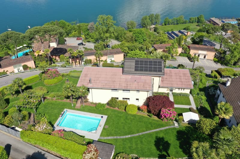 Aerial view of a large, two-story house with a swimming pool, surrounded by lush greenery, gardens, and palm trees. The house has solar panels installed on the roof. The property is situated on a hillside overlooking a body of water, likely a lake or rive