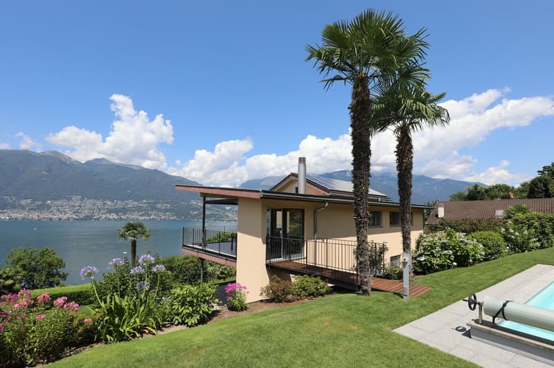 Two-story house with a balcony, surrounded by a well-maintained garden with palm trees and other plants. The house overlooks a lake or body of water, with mountains in the background. There is also a swimming pool visible in the image.
