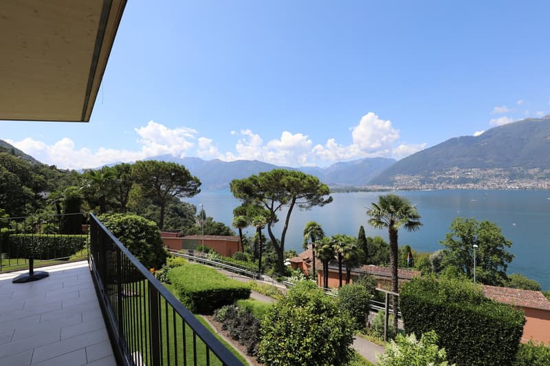 The image shows a balcony or terrace overlooking a scenic lake surrounded by mountains and lush vegetation. The balcony has a metal railing and the view includes palm trees, pine trees, and other greenery. The lake and mountains can be seen in the distanc