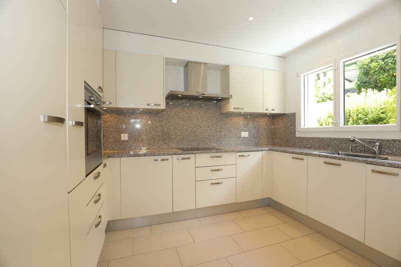 Spacious modern kitchen with beige cabinets, granite countertops, stainless steel appliances including a dishwasher, and large windows overlooking the greenery outside.