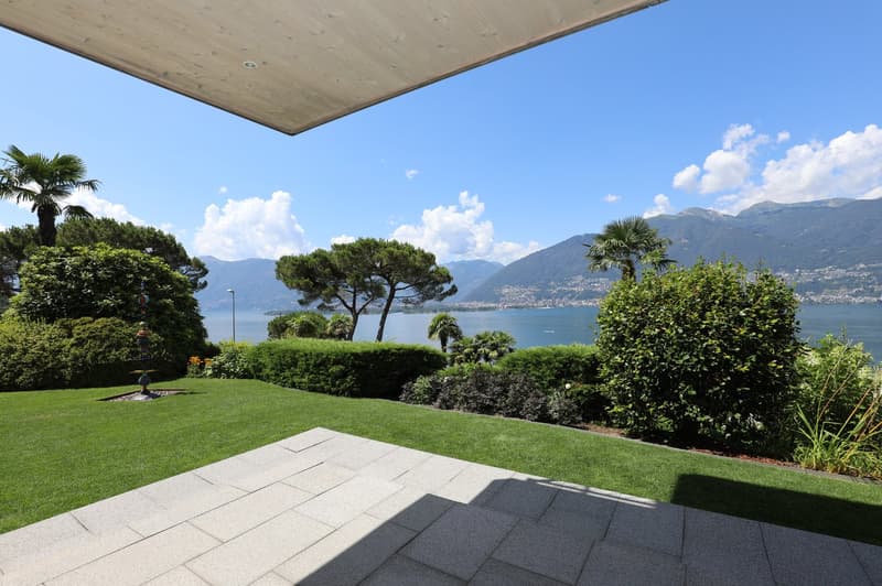 Spacious outdoor terrace with a panoramic view of a lake surrounded by mountains and lush greenery, including palm trees and other tropical plants. The terrace has a wooden ceiling and a well-manicured lawn with a fountain.