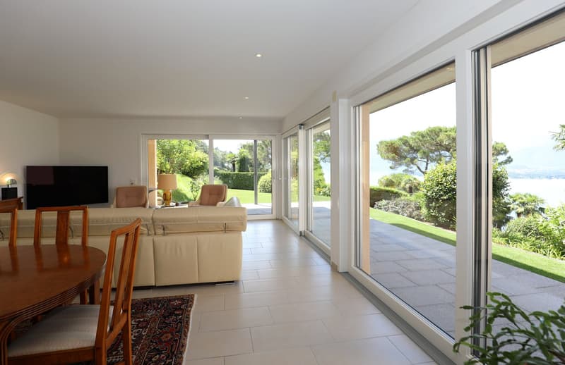 Spacious living room with large windows overlooking a lush garden and scenic view, hardwood dining table, comfortable sofa set, and tiled flooring