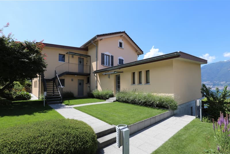 2-story house with a balcony, yellow exterior, red roof, surrounded by a well-maintained lawn and landscaping, with stairs leading up to the entrance.
