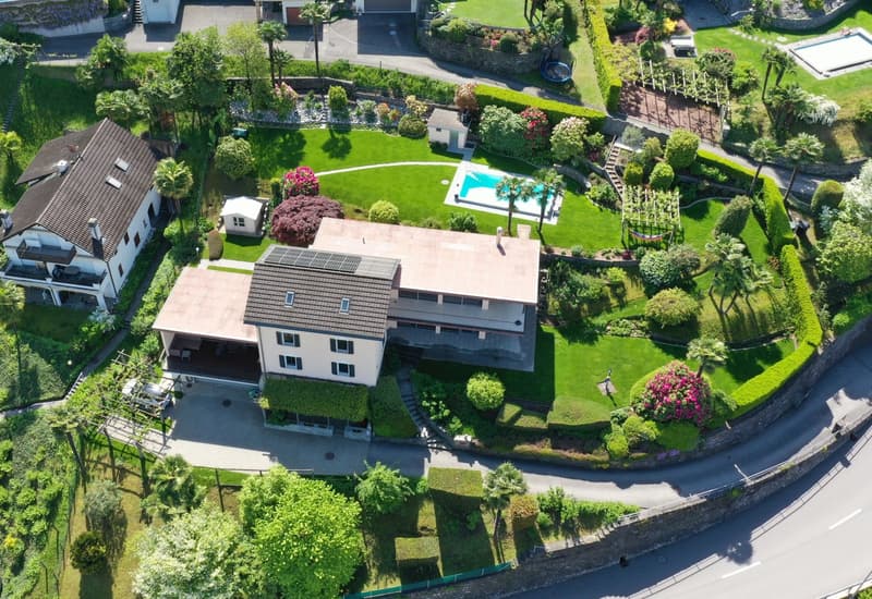 Two-story house with a red tile roof, surrounded by a well-manicured garden with a swimming pool, lush greenery, and various landscaping features such as hedges and flower beds.