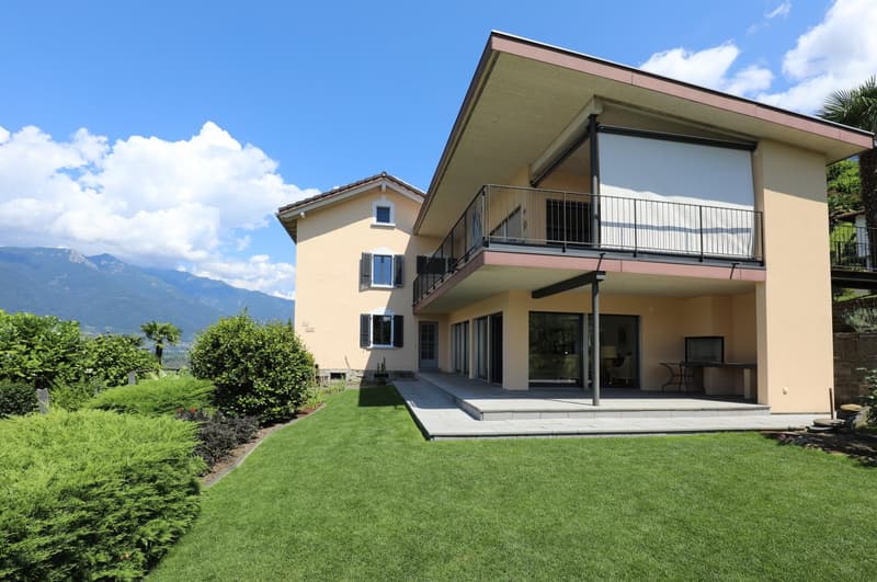 2-story house with a yellow exterior, large balcony, and a well-maintained lawn and garden. The house has a modern design with large windows and sliding glass doors leading to the outdoor patio area. The surrounding landscape includes mountains and lush g