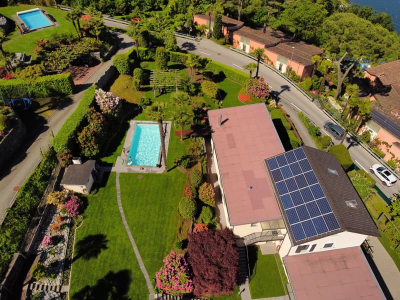 Aerial view of a large, two-story house with a red tile roof, surrounded by a well-manicured garden with a swimming pool, solar panels on the roof, and a parking area.