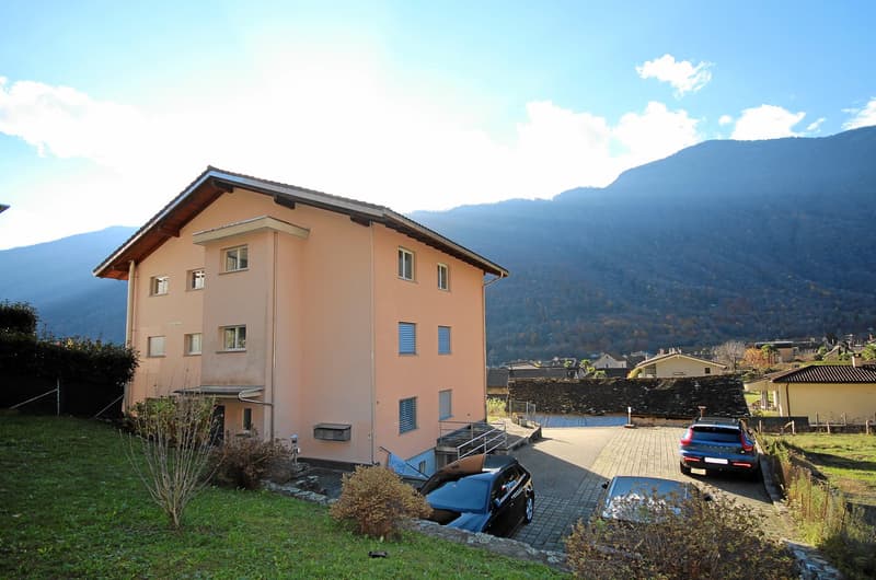 2-story house with beige exterior, several cars parked in the driveway, green grass, and mountains in the background.