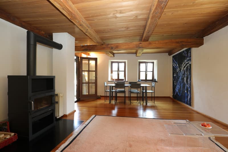 Spacious living room with wooden beamed ceiling, large windows, wood-burning stove, dining table and chairs, and a decorative artwork on the wall