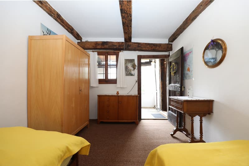 Spacious bedroom with exposed wooden beams, wooden furniture including a wardrobe and dresser, and a fireplace. The room has a cozy and rustic feel with yellow bedding and a wooden floor.