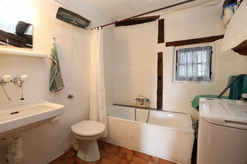 Bathroom with a bathtub, toilet, sink, and a private washing machine. The room has white walls, wooden beams, and a small window with checkered curtains.