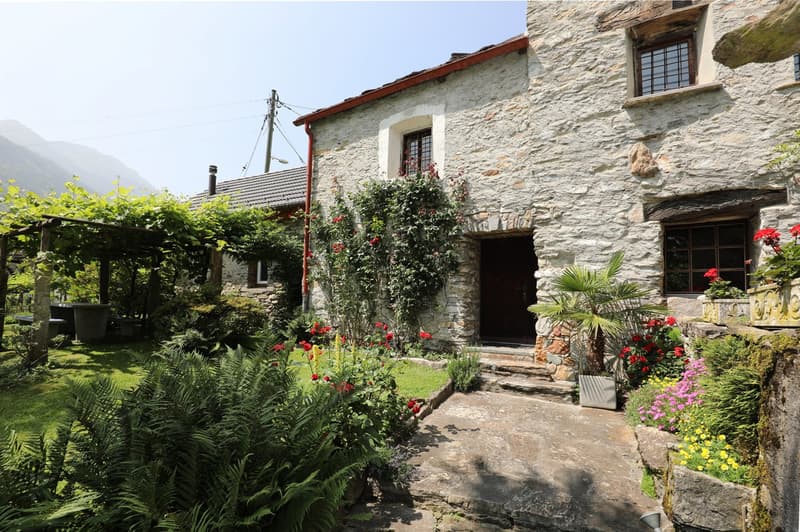 Two-story stone house with a red tile roof, surrounded by a lush garden with various flowering plants and a palm tree. The house has a covered patio or balcony area, and there appears to be a chimney or fireplace visible. The entrance to the house is acce