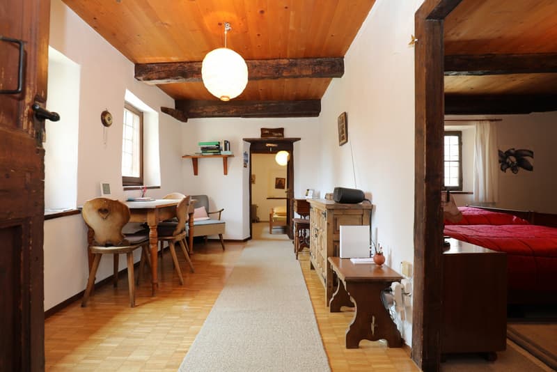 Cozy rustic living space with wooden beamed ceiling, wooden furniture, and a fireplace. The room has a dining area with a wooden table and chairs, as well as a seating area with a red sofa. The space also includes shelves, a cabinet, and other decorative