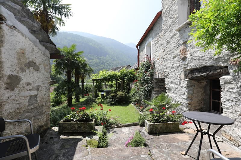 A charming stone house surrounded by a lush garden with palm trees, flowers, and a patio area with a table and chairs. The house is situated in a scenic mountainous landscape with green hills in the background.