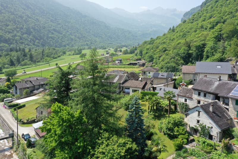 The image shows a picturesque mountain village nestled in a lush, green valley. The village is surrounded by densely forested hills and mountains, creating a serene and tranquil atmosphere. The buildings in the village have traditional alpine architecture