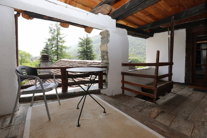 Covered outdoor patio with wooden beams, stone walls, and a table with chairs overlooking a forested mountain landscape