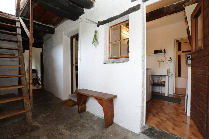 This image shows the interior of a rustic, traditional-style room. The walls are white with exposed wooden beams and a wooden bench along one wall. There is a small window with a wooden frame, and a hanging plant decoration. The floor appears to be made o