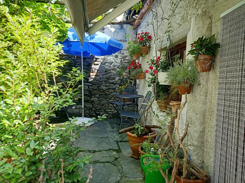 Small outdoor patio area with stone walls, potted plants, a blue umbrella, and a table with chairs. The space is surrounded by lush greenery and has a cozy, rustic feel.