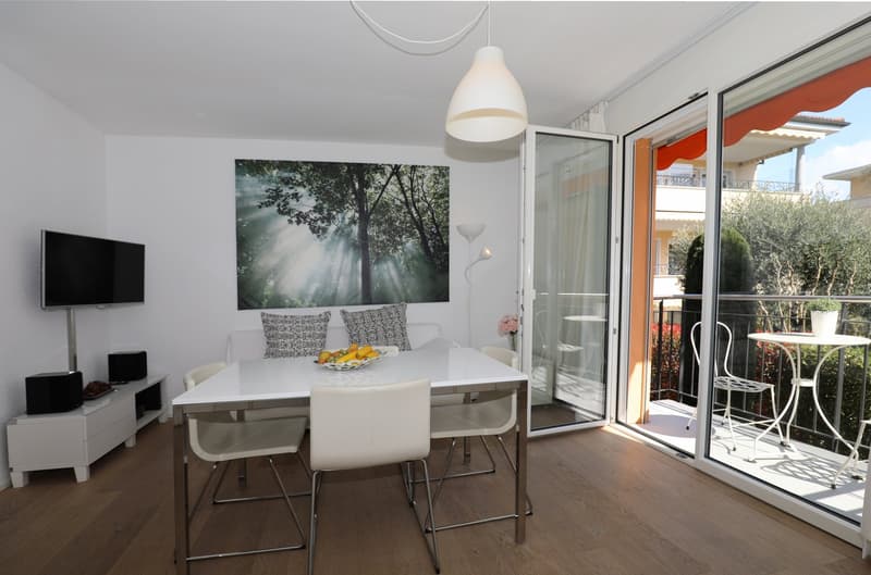 Dining area with a white table and chairs, a white couch with pillows, a TV, a white cabinet, and a lamp. There is a balcony with a table and chairs.