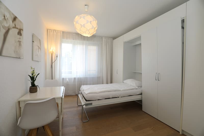 small room with bed, desk, chair, lamp, and flower vase