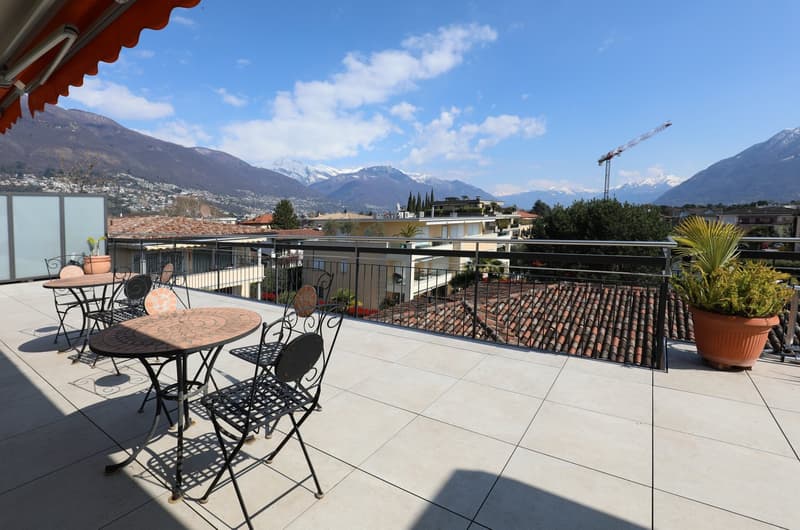 Outdoor balcony with tables and chairs, metal railing, potted plants, tiled floor, view of mountains and city