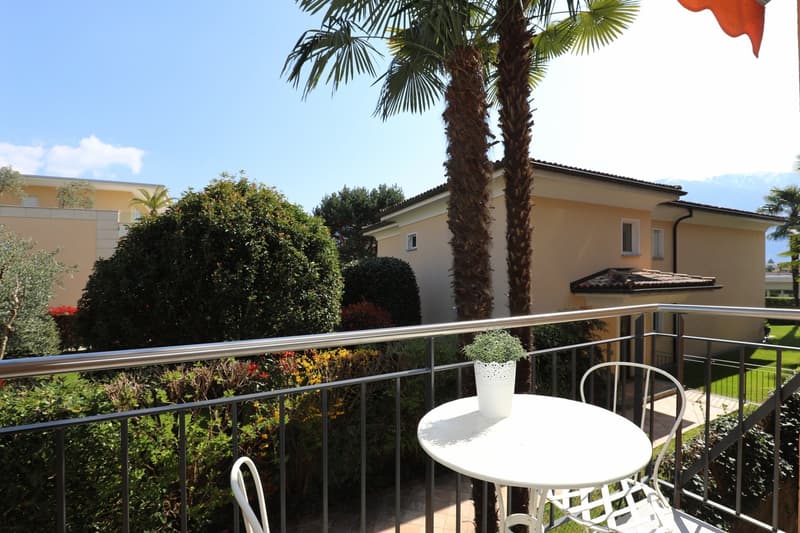 Balcony with metal railing, white table and chairs, potted plant, view of a house and garden