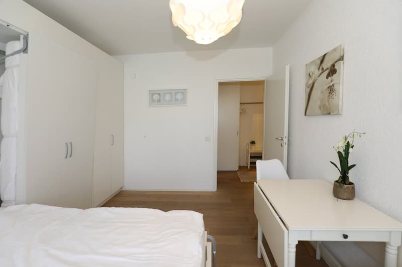 Bedroom with bed, desk, white wardrobe, white walls, wooden floor, lamp, picture frame, and a chair.
