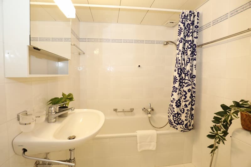Bathroom with white tiles, white sink, white bathtub, shower curtain, potted plants, towel on bathtub, white toilet