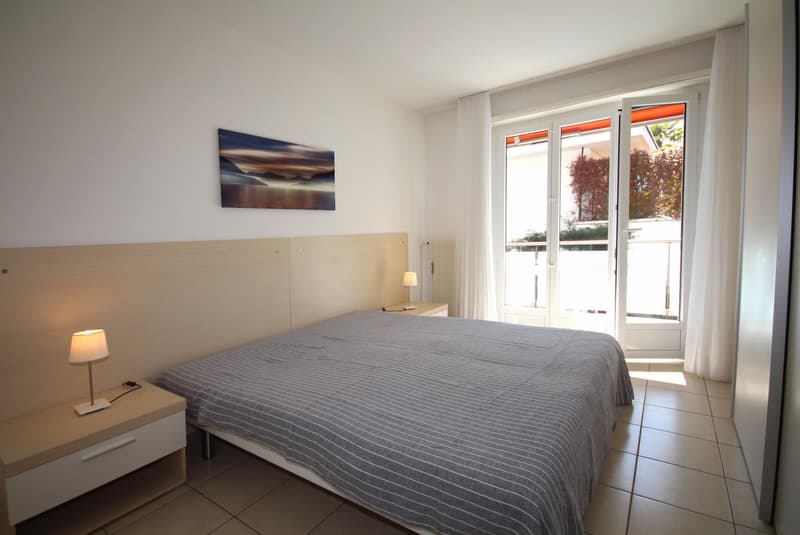 A bedroom with a bed, nightstands, lamps, and a painting on the wall. There is a balcony with glass doors.