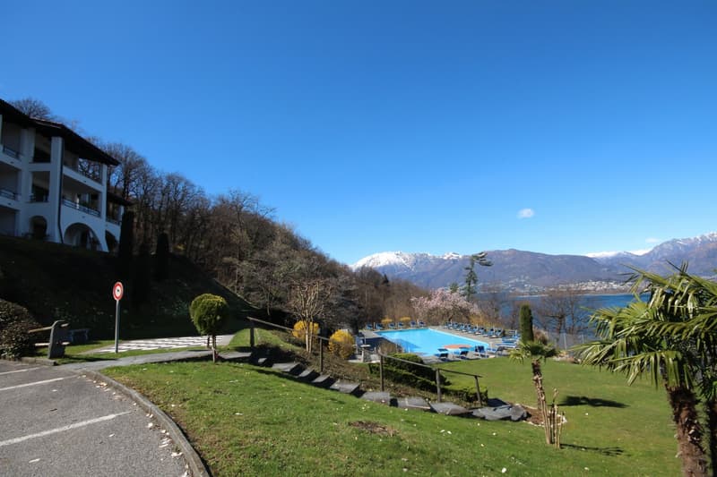 The image shows a hotel or resort property situated on a hillside overlooking a lake. The building has a classic European architectural style with balconies and arched windows. The property has a large outdoor swimming pool surrounded by lush greenery and