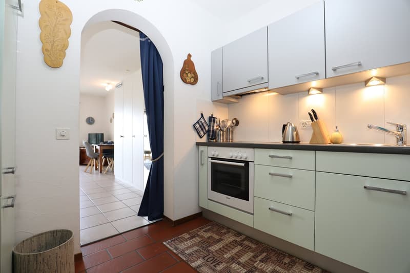 Spacious kitchen with white cabinets, black countertops, built-in oven, and various kitchen utensils and appliances. The kitchen leads to an adjacent room through an arched doorway.