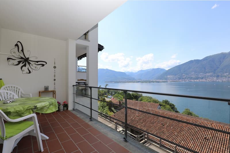 Bedroom with a balcony overlooking a lake and mountains. The room has white walls, a tiled floor, and a large window with a view of the scenic landscape outside.