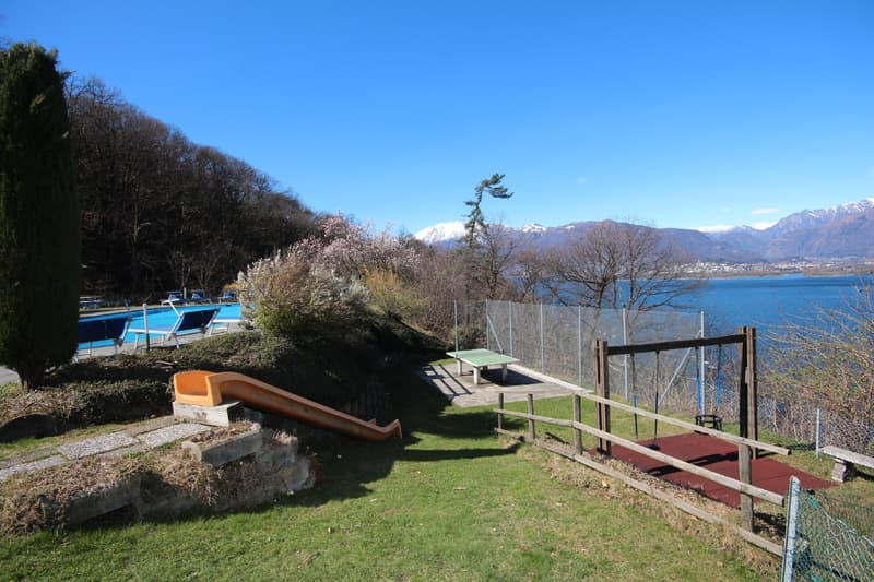 Outdoor area with a swimming pool, benches, and a view of the lake and mountains in the background. The area is surrounded by trees and has a grassy lawn.