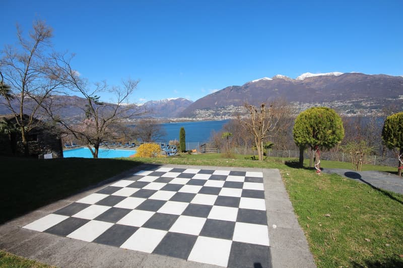 Outdoor area with a checkerboard-patterned patio, surrounded by trees and shrubs, overlooking a lake or body of water with mountains in the background. There is also a swimming pool visible in the image.