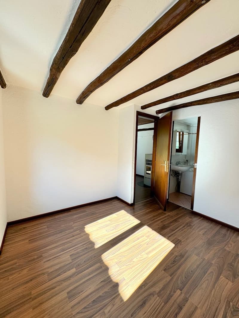 The image shows a room with exposed wooden beams on the ceiling, hardwood flooring, and a doorway leading to another room. The room appears to be a living or bedroom space with natural light coming in through the windows.