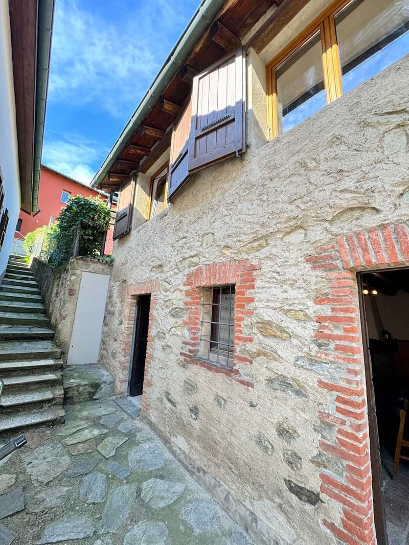 The image shows the exterior of an old stone building with wooden beams and shuttered windows. The building has a stone facade with some brick accents, and there is a set of stairs leading up to the entrance. The surrounding area appears to be a quaint, h
