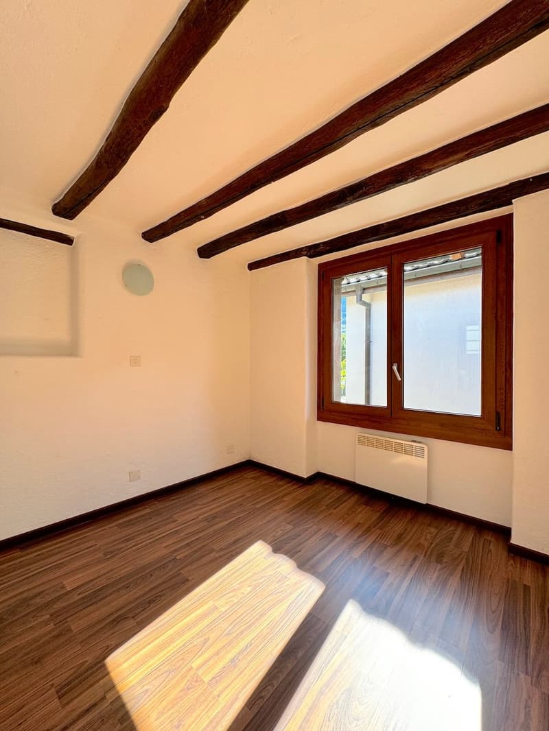The image shows a room with wooden beams on the ceiling, a wooden window frame, and a hardwood floor. The walls are painted a light color, and the room appears to be well-lit with natural light coming in through the window.